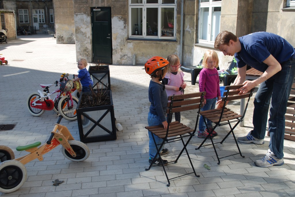 3 børn inspicerer en fars arbejde med stolene, mens et andet barn knokler med en cykel i baggrunden.