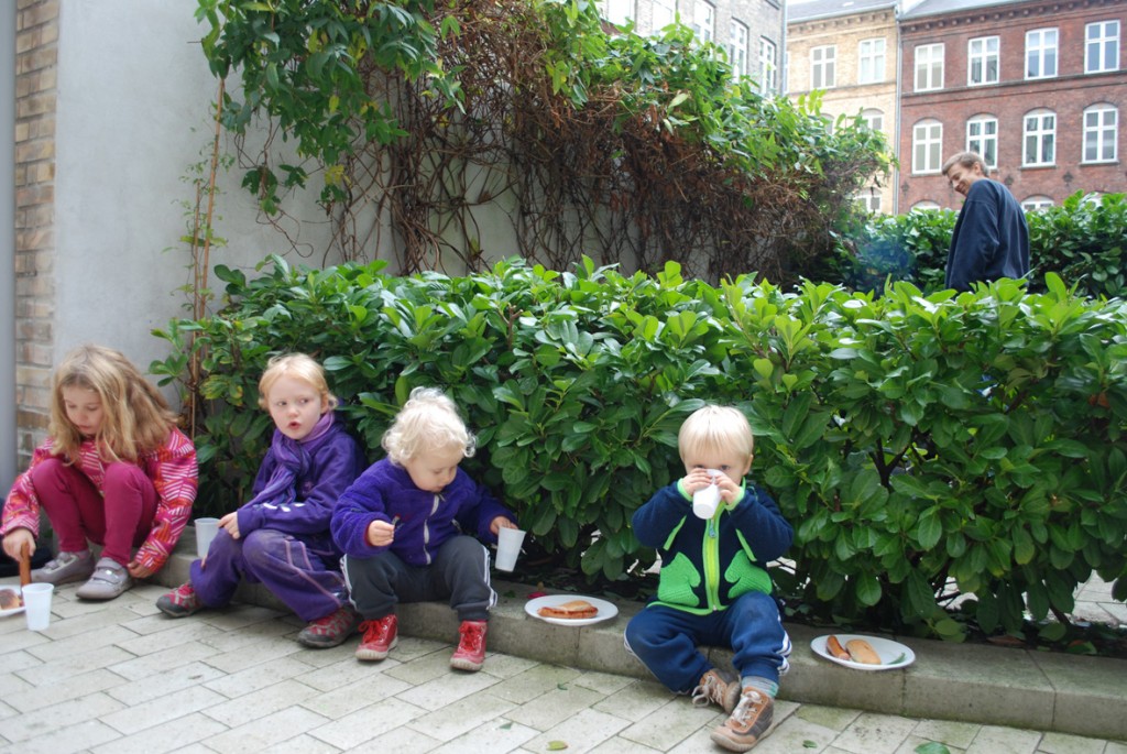 4 børn nyder pølser med brød og saft på kantstenen i gården.